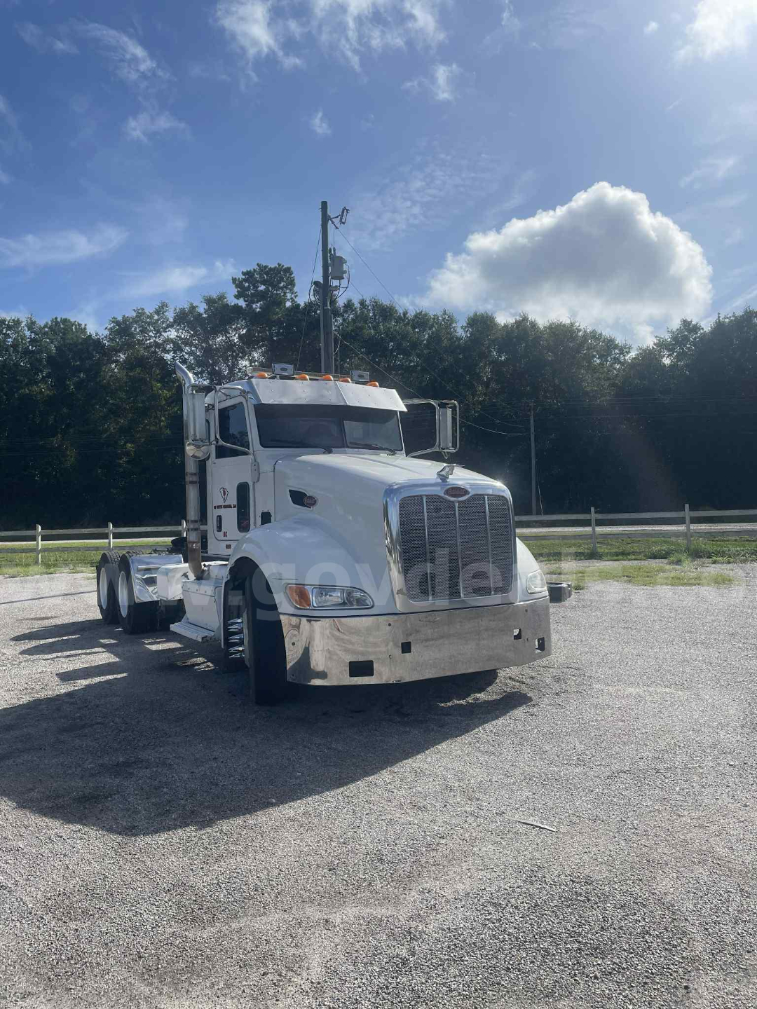 White 2012 Peterbilt 386 truck cab and chassis with chrome front bumper and large grille, single sleeper, orange clearance lights on roof, dual rear axles, parked on gravel.