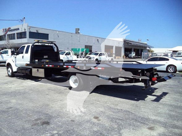 Tow truck with flatbed trailer attached, white cab, stock number T26834FM visible in a parking lot with multiple vehicles and a large industrial building in the background.
