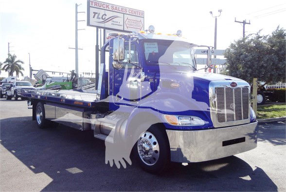 Blue Peterbilt 337 tow truck with chrome front bumper and flatbed, dealership lot in background.
