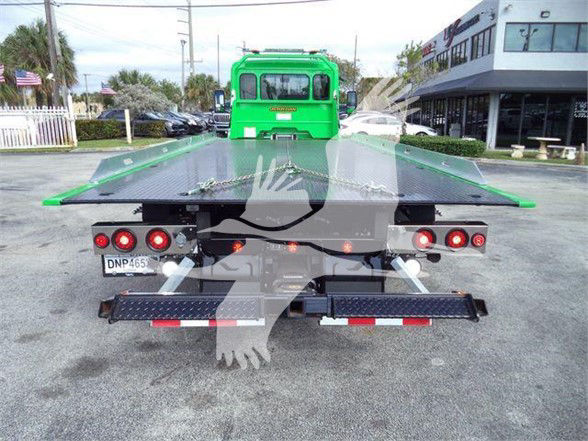 Flatbed tow truck rear view with green cab, platform bed, multiple lights, and license plate visible.