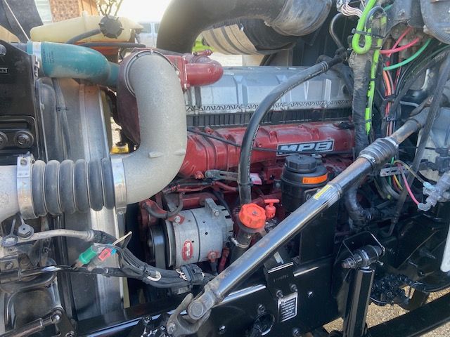 Engine bay of a Mack CXU613 showing MBP engine with red valve cover, hoses, wiring harness, and various components.