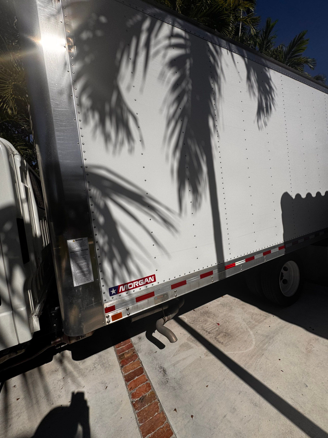 Morgan box truck trailer with white cargo area and reflective red-white safety stripe showing Morgan branding. Side panel with riveted aluminum and shadow of palm trees.