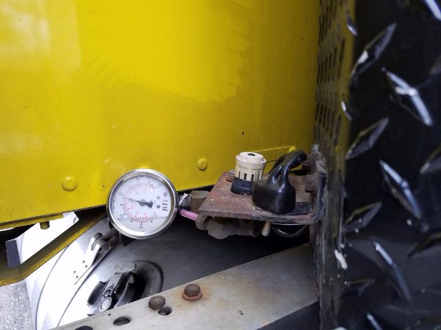Gauge gauge and fittings mounted on yellow truck body with black diamond plate tread. A round pressure gauge shows numbers, connected to a small brown metal bracket with a knob and plug.