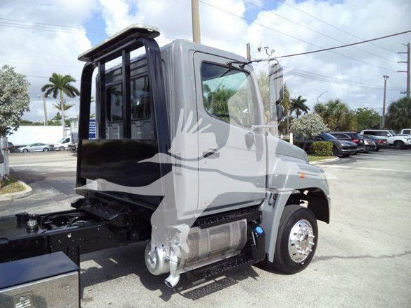 Truck cab of a 2014 Hino 258ALP in a dealership lot showing front fender, door, and side step with no engine cover. The image includes watermark overlay of a stylized hand.