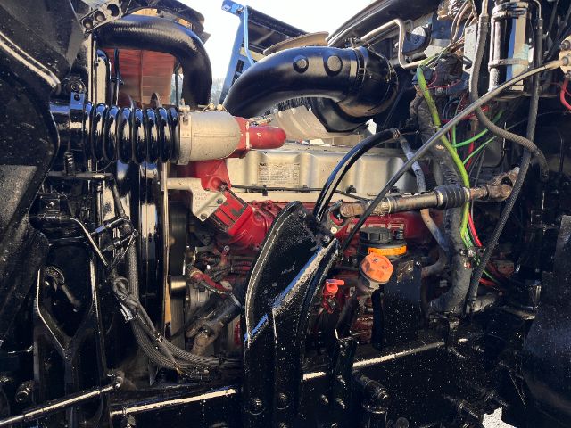 Engine bay of 2013 Mack CHU613 Pinnacle showing coolant hoses, red engine components, black air intake, orange dipstick handle, wiring, and metal framework.