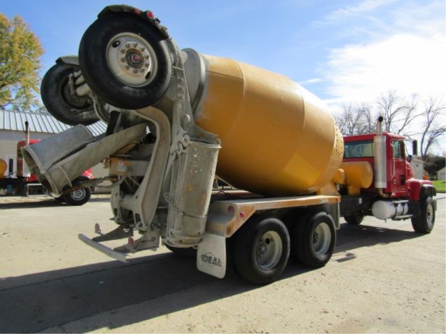 Concrete mixer truck with drum tilted sideways on trailer attached to red cab; spare wheel mounted on mixer, outdoor yard setting.