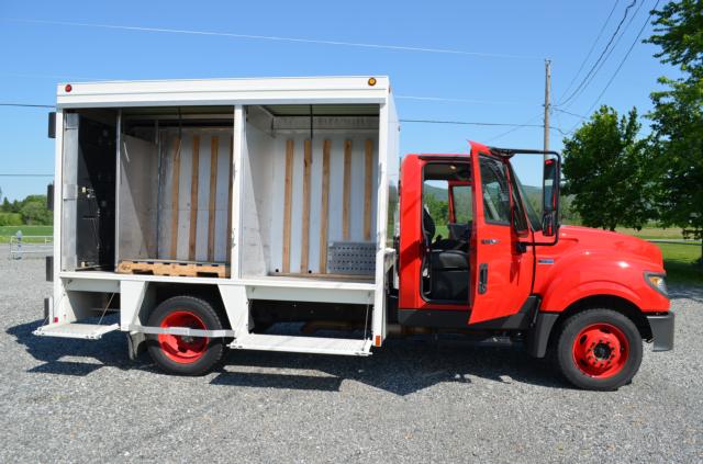 Truck side view: red International TerraStar medium-duty truck with white box cargo body, open rear and side doors, empty wooden framing inside cargo area.