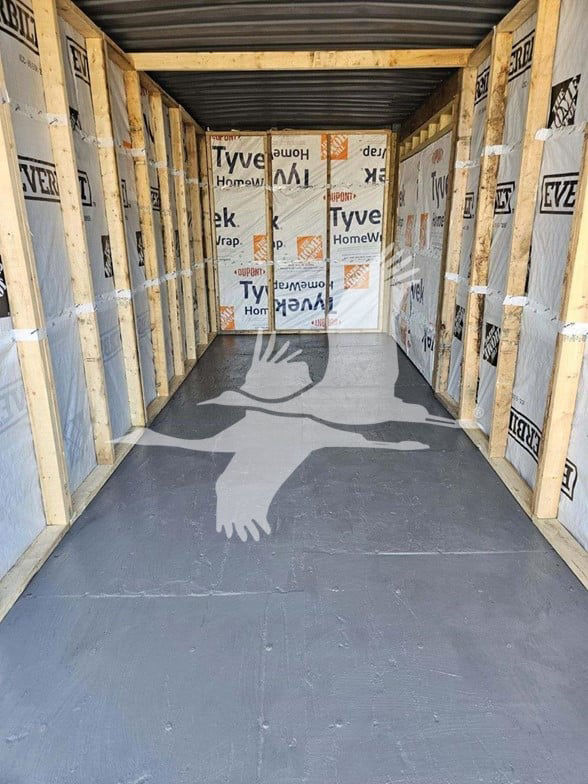 Trailer interior with wooden studs and Tyvek insulation, finished dark floor panels; rectangular space resembling a container being converted; visible studs on both sides.