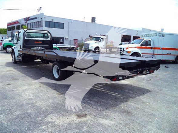 Flatbed tow truck with extended long bed on a Freightliner chassis, service yard with buildings in background.