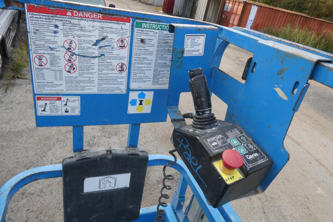 Truck-mounted Genie GS1930 scissor lift control panel and safety labels on blue platform, joystick, emergency stop button, and warning placards.