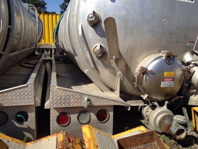 Vacuum tank trailer showing rear side with circular manway hatch, welded aluminum body, trailing axles, and hazard label on access cap.