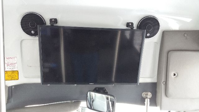 Vehicle interior dashboard with a mounted flat-screen monitor between two circular speakers. Left shows a small warning sticker, right has a panel. Mirror at bottom. No engine text visible.