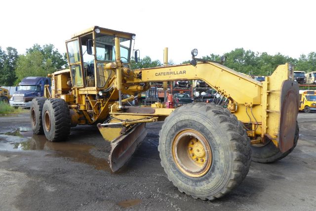 Caterpillar 16G motor grader in a yard showing large front wheel and front moldboard with operator cab, yellow paint.