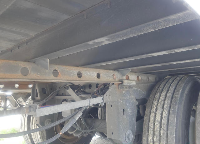 Trailer undercarriage showing frame with perforated crossmember, electrical wiring, suspension components, and dual tires. Rusty metal surfaces and a hose/cord routing along the frame.