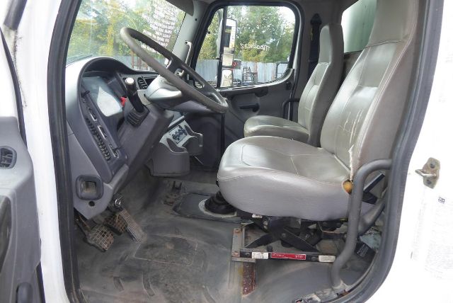 Freightliner M2 106 cab interior showing steering wheel, dashboard, and two gray cloth seats with floor mats.