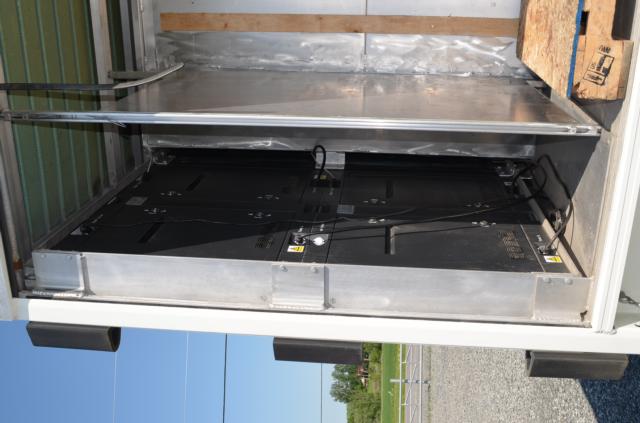 Truck cargo area showing interior of a rear compartment with a large electronic console or panel mounted inside, some cables, metal framing, and a wooden pallet on the floor.