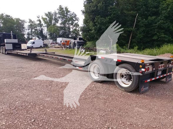 Trailer showing long lowboy with dual axles at rear, flat deck, and wheel wells. Visible text on rear: no legible brand or serial from distance. VOI/stock area not clearly readable.