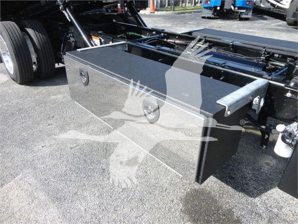 Flatbed area of a 2023 International MV truck showing a large rectangular metal toolbox mounted behind the cab with latches on the front and side, and part of the chassis and wheels visible.