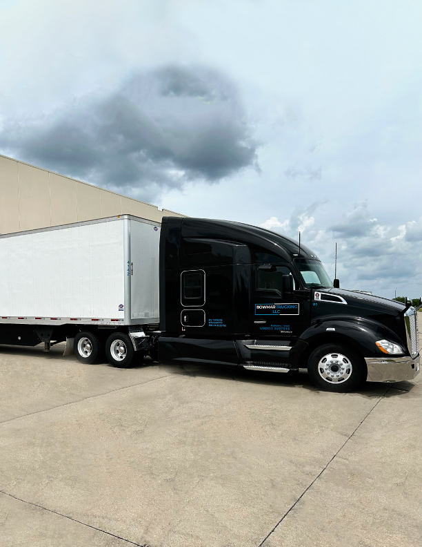 Kenworth T680 tractor attached to a white trailer in a parking lot, black cab with logo on door, chrome steps and wheels visible.