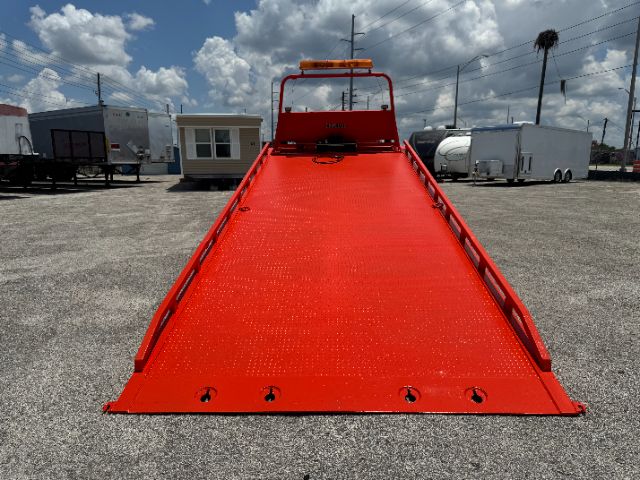 Tow truck with bright red flatbed ramp, loading system, and safety rails. Flatbed surface shows textured metal with tie-down points along edges. Tow truck cab not visible.