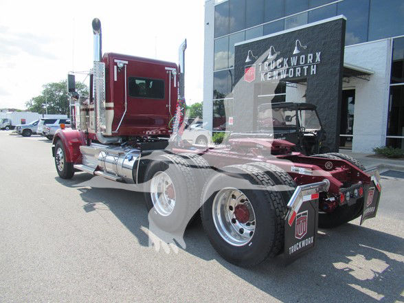 Kenworth W900L conventional daycab truck with maroon cab and polished exhaust stacks, chrome fuel tanks, and dual rear axles in front of a truck dealership.