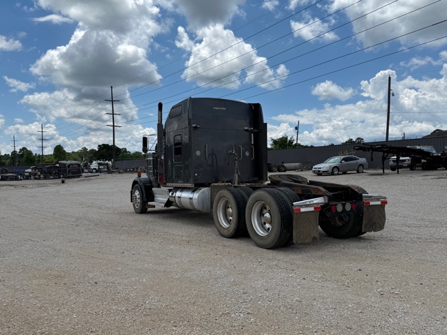 Kenworth W900 semi-truck tractor in a gravel lot from rear three-quarter view showing sleeper cab and dual rear axles with flatbed trailer hitch.