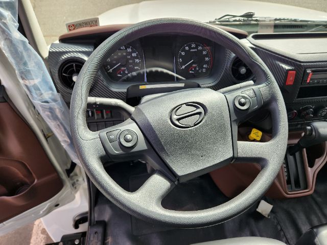 Steering wheel of a Hino truck cabin with dashboard controls visible. View includes instrument cluster with speedometer and tachometer, left stalk controls, and right side dash.