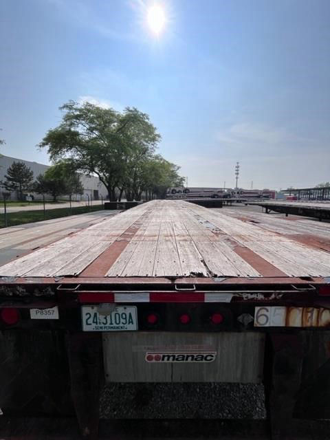Flatbed trailer deck with wooden planks and red steel crossmembers, Manac logo on rear plate area, license plate visible.