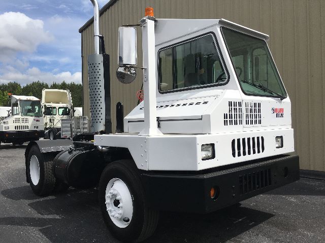 Kalmar Ottawa YT30 yard truck chassis with white cab and black bumper, orange beacon on roof, side exhaust, and multiple similar trucks in background.