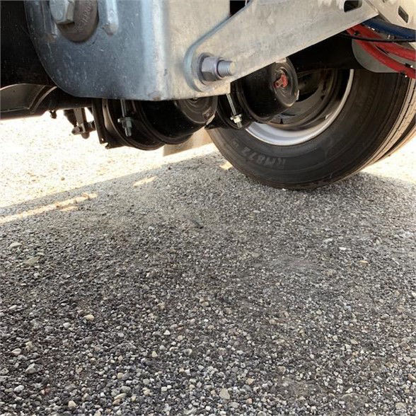 Flatbed trailer undercarriage showing wheel, suspension leaf springs, and frame. Visible tire with tread and small bolts on suspension mount.