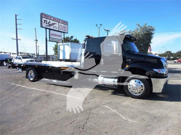 Flatbed tow truck (black) parked outdoors with chrome wheels and a silver flatbed, showroom-like setting, signboards in background.