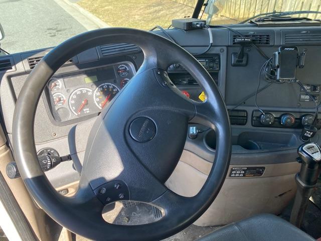 Dashboard and steering wheel of a Freightliner Cascadia cab interior. Visible gauges, steering wheel center horn, gear shifter, control panels, and mounted devices.