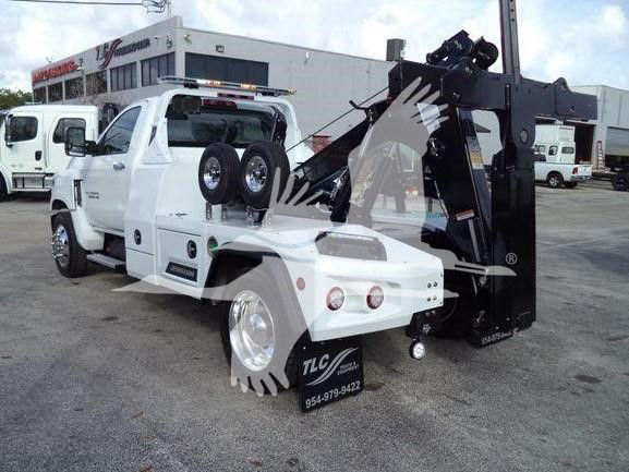 Tow truck with a large black hydraulic lift behind cab; white Silverado 6500HD tow bed; spare tires mounted on side; dealership lot visible in background; TLC logo and contact number on front bumper.