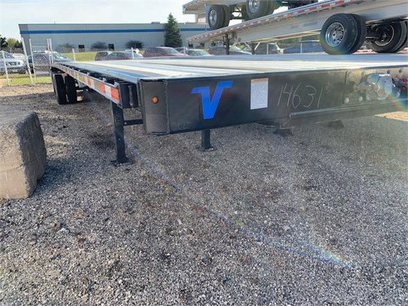 2024 Vanguard steel flatbed trailer front-left side showing V logo and side rails with a V-shaped support and gravel ground.