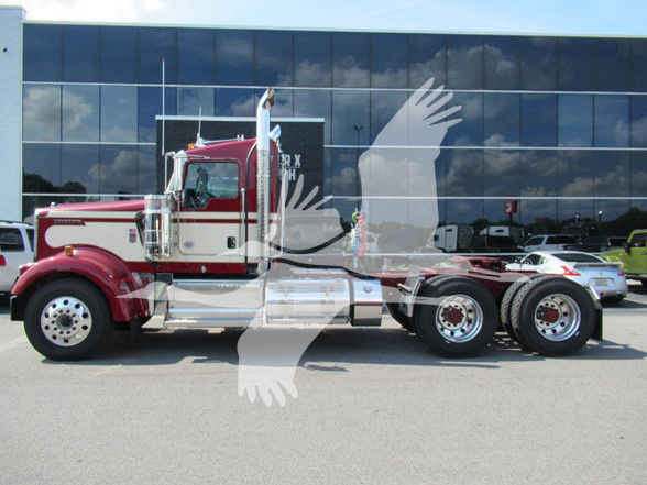 Kenworth W900L conventional daycab truck in red and chrome, side profile showing dual rear axles and chrome fuel tanks; showroom building reflected in glass facade.