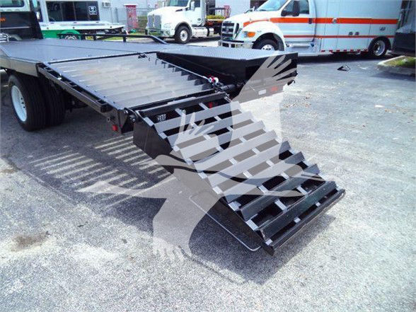 Flatbed truck trailer with ramp and steps, rear view showing loading ramp extending from black flatbed. Ambulance and other trucks in background.