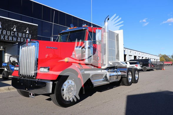 Kenworth W900L truck in red with chrome exhaust stacks and large grille, parked in front of dealership.