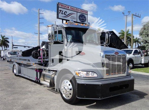 Peterbilt 337 tow truck front quarter view with tilt bed and chrome grille, silver cab, black bumper, and a signboard in background.