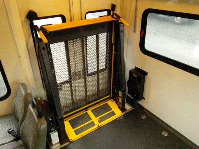 Ford E-450 bus interior with wheelchair lift folded up against wall, yellow safety rails, mesh gate, and seating area.