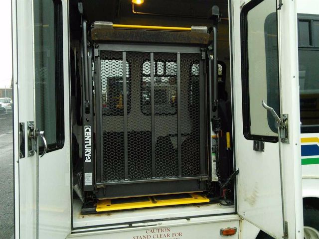 Bus rear interior showing a wheelchair lift with metal mesh gate and yellow platform, doorway open, surrounding white bus with door frame and caution label at bottom.