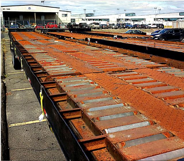 Rolloff trailer bed with rusted orange steel components and multiple tracks, parking lot in background with buildings.