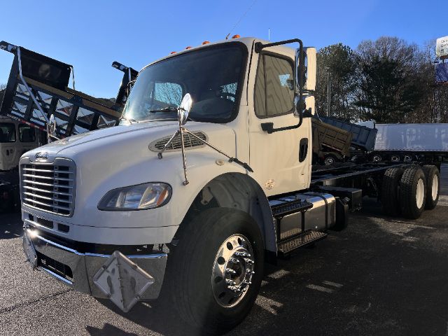 Truck cab and chassis showing Freightliner M2 badge, white exterior, large side mirrors, dual rear wheels, front bumper with protective diamond plate and chrome hubcaps.