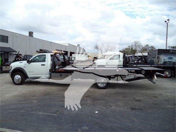 Ram 5500 tow truck with flatbed, white cab, dealership lot in background, VIN and stock number visible but not legible details.