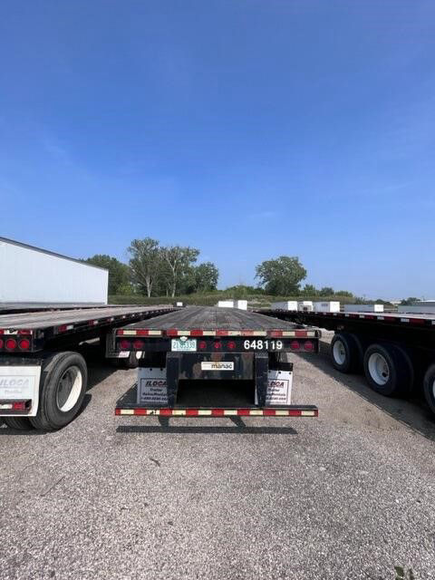 Flatbed trailer with Manac branding and stock numbers 648119 and 80482 visible on the rear plate area. Open space background with blue sky.