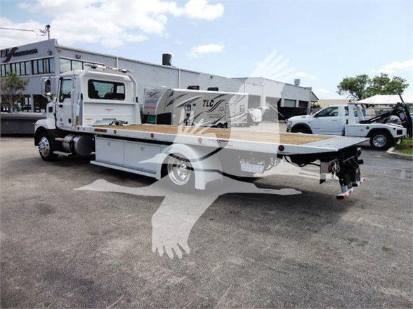 Flatbed tow truck with a white cab and long flatbed, parked in a dealership lot. There are buildings and other trucks in the background.