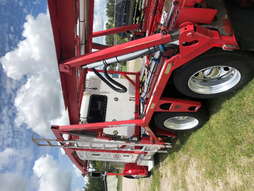 Peterbilt 379 auto carrier truck side view showing white sleeper cab and red car-hauler frame with hydraulic components and dual rear axles.