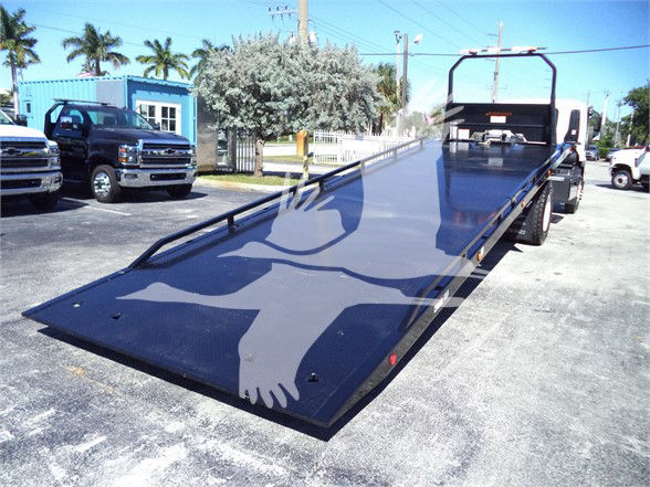 Isuzu FTR flatbed tow truck with a long ramp, rear loading platform, and wheel lift; no visible text on the ramp, dealership yard in background.