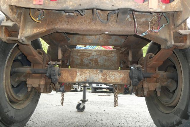 Rear view of a Fruehauf trailer axle assembly showing rusted steel frame, dual axles, leaf springs, and central hitch area with chains. Various wiring and a support jack visible.