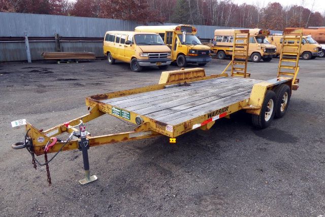 Trailers in a yard: a Warrior tilt-bed trailer with dual rear ramps, metal frame, wood deck, hitch and safety chains; visible wheels and front jack stand.