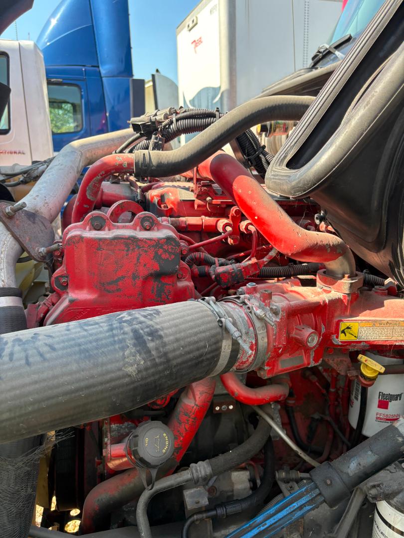 Red engine bay of a 2016 International Prostar with multiple hoses and a large black intake tube visible. Various red components, yellow warning label, and Fleetguard filter visible.